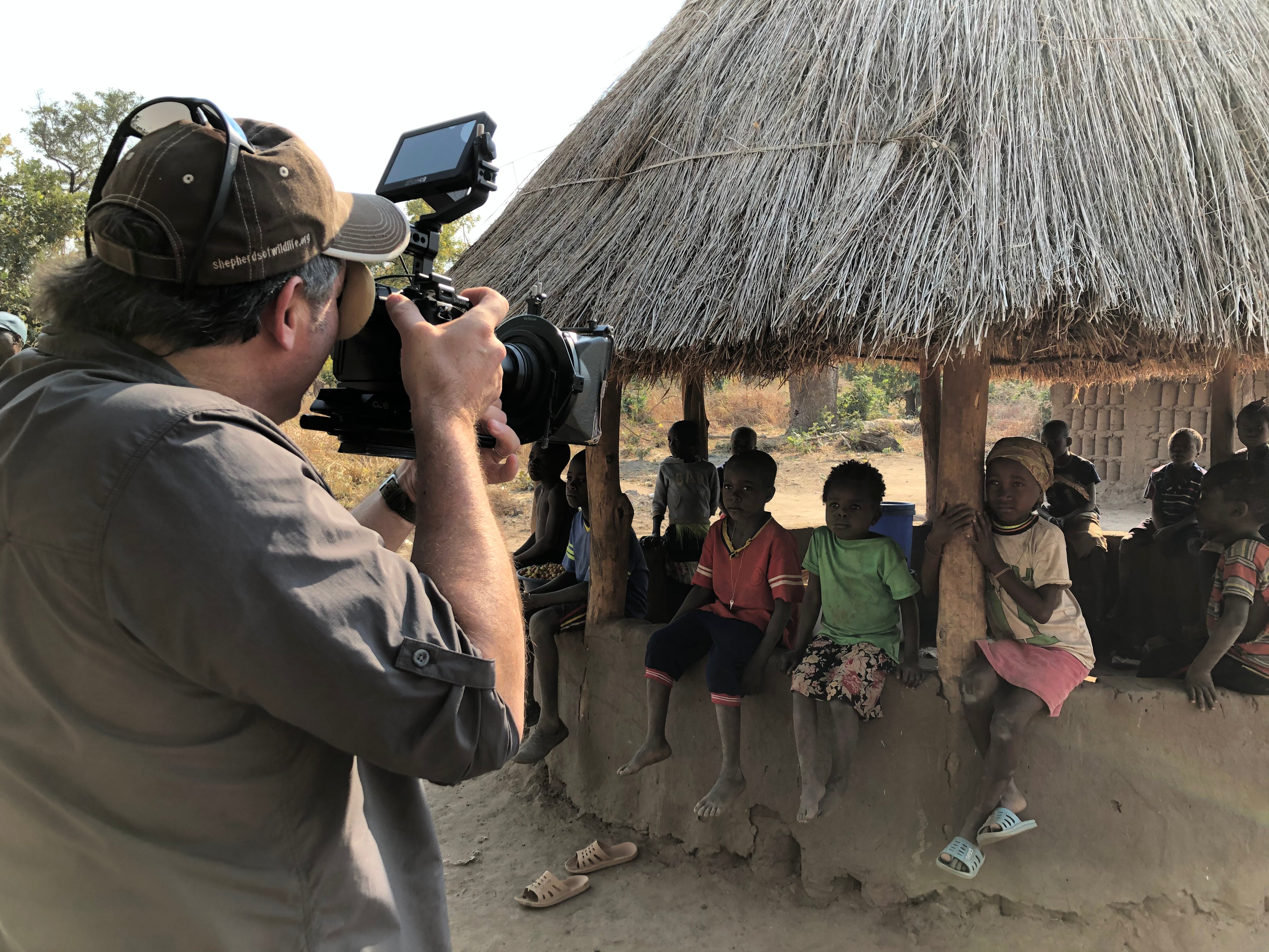 Person filming a group of children in front of a thatched-roof structure.