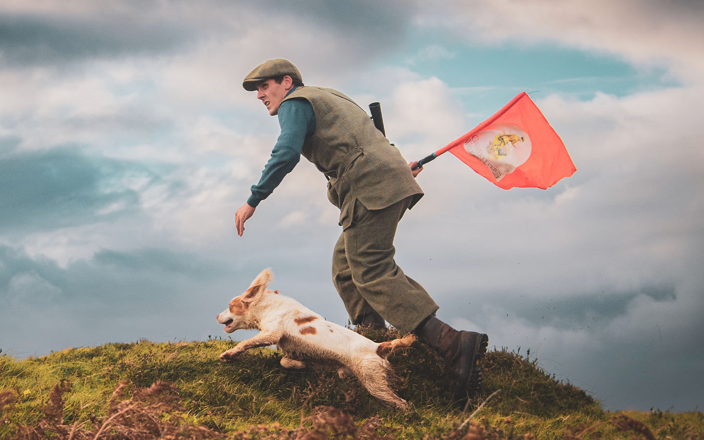 The Last Keeper - Scottish Man in hunting attire with a dog on a grassy hill, holding a red flag with a logo.