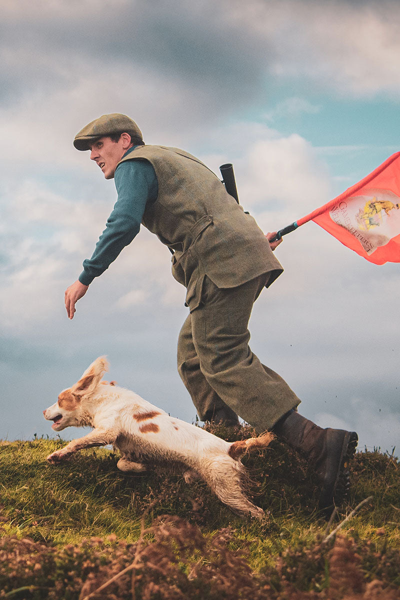 The Last Keeper - Scottish Man in hunting attire with a dog on a grassy hill, holding a red flag with a logo.