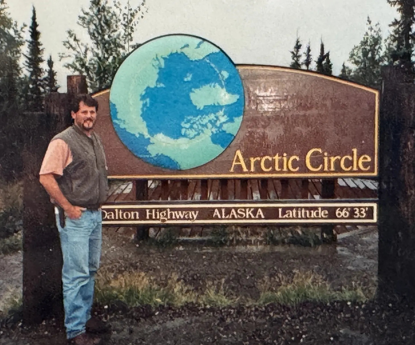 Tom Opre stands beside a large wooden sign reading 'Arctic Circle' with a blue-and-white globe graphic and text indicating Dalton Highway, Alaska, Latitude 66 33'. Trees and an overcast sky are visible in the background.